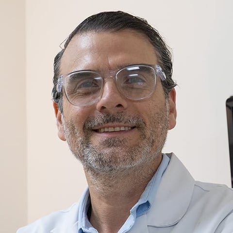 Middle-aged man with gray beard wearing clear-framed glasses and light blue shirt, smiling at camera