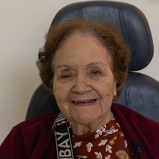 Elderly woman with reddish-brown hair wearing a burgundy cardigan over a patterned brown top, seated in a dark office chair, smiling warmly at the camera